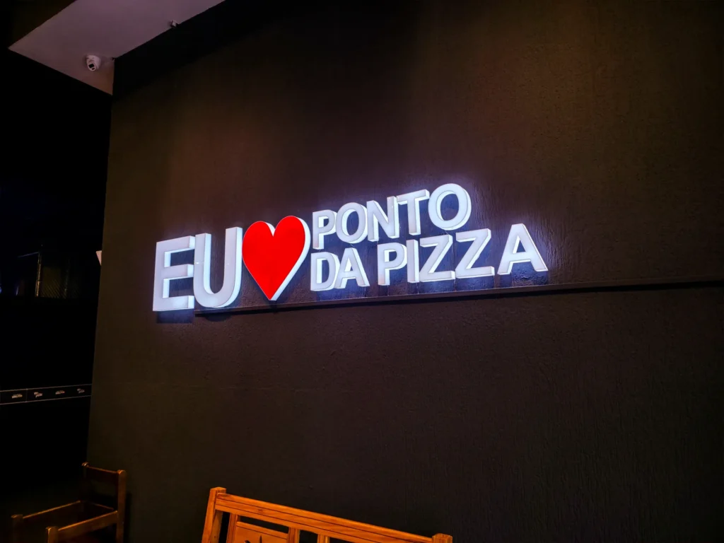 Frase luminosa Eu amo Ponto da Pizza em parede escura, com um banco de madeira à frente